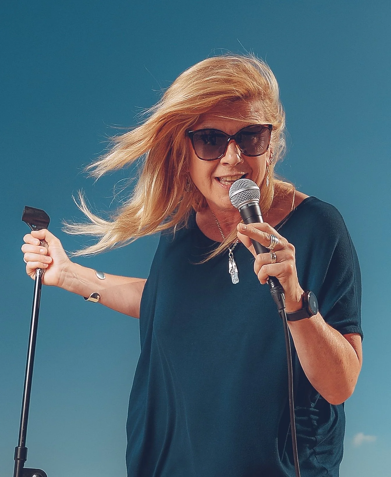 A woman with blonde hair wearing sunglasses, a black shirt, holding a microphone and a cane, singing against a clear blue sky.