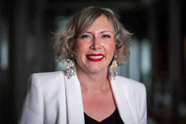 A woman with short, curly gray hair wearing a white blazer and large, ornate earrings, smiling in an indoor setting.