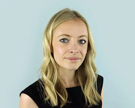Portrait of a young woman with blonde hair and light skin, wearing a black sleeveless top, standing against a light blue background.