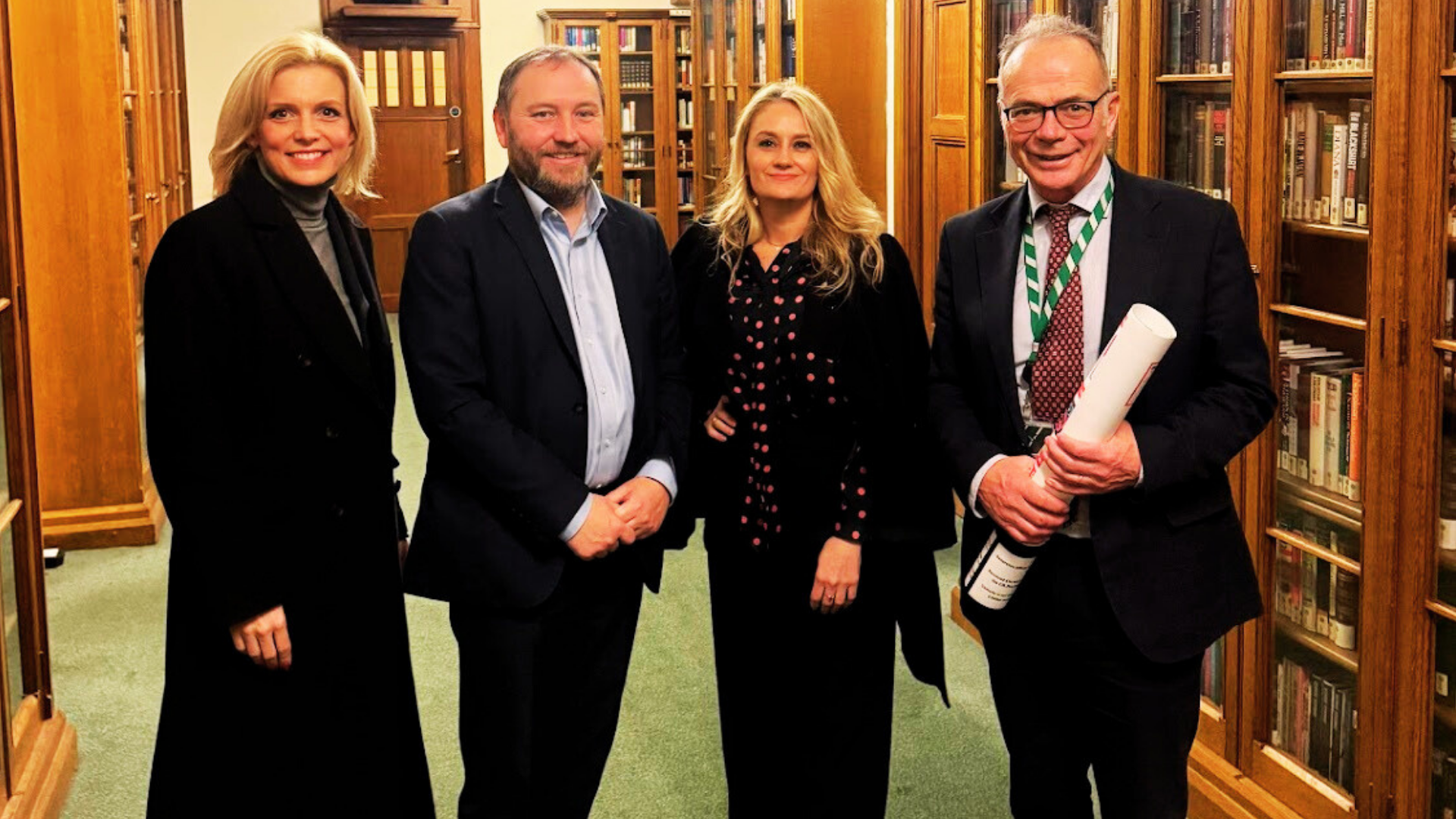 Four people standing in a library with wooden bookshelves. The group includes two women and two men, all dressed in formal attire, smiling at the camera. One man is holding a rolled-up document.