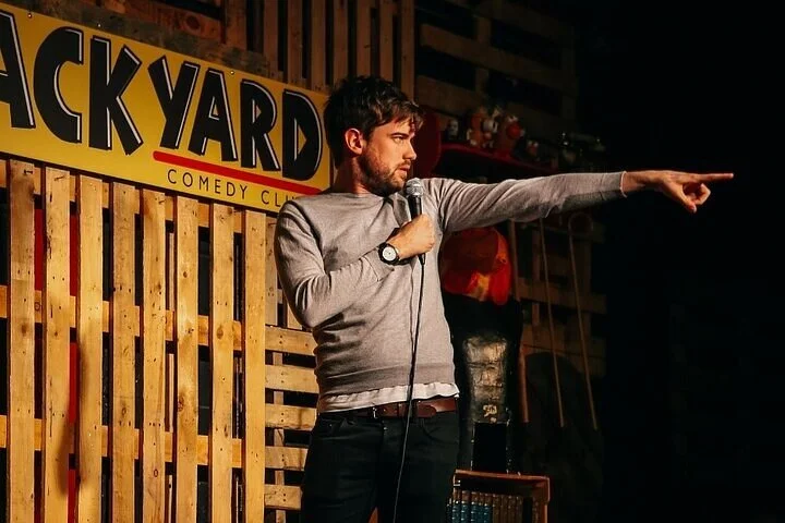 A male comedian with beard and medium-length hair stands on stage at a comedy club, holding a microphone in his right hand, and extending his left arm, pointing to the right. He is wearing a gray long-sleeve shirt with white underneath and a watch on his left wrist. The background features a wooden wall with a yellow sign that reads 'BACKYARD COMEDY CLUB' in black and red letters.