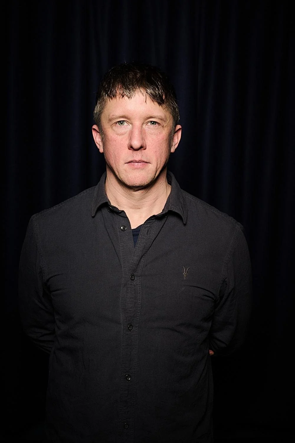 A man with light skin, short dark hair wearing a black button-up shirt standing in front of a dark curtain.