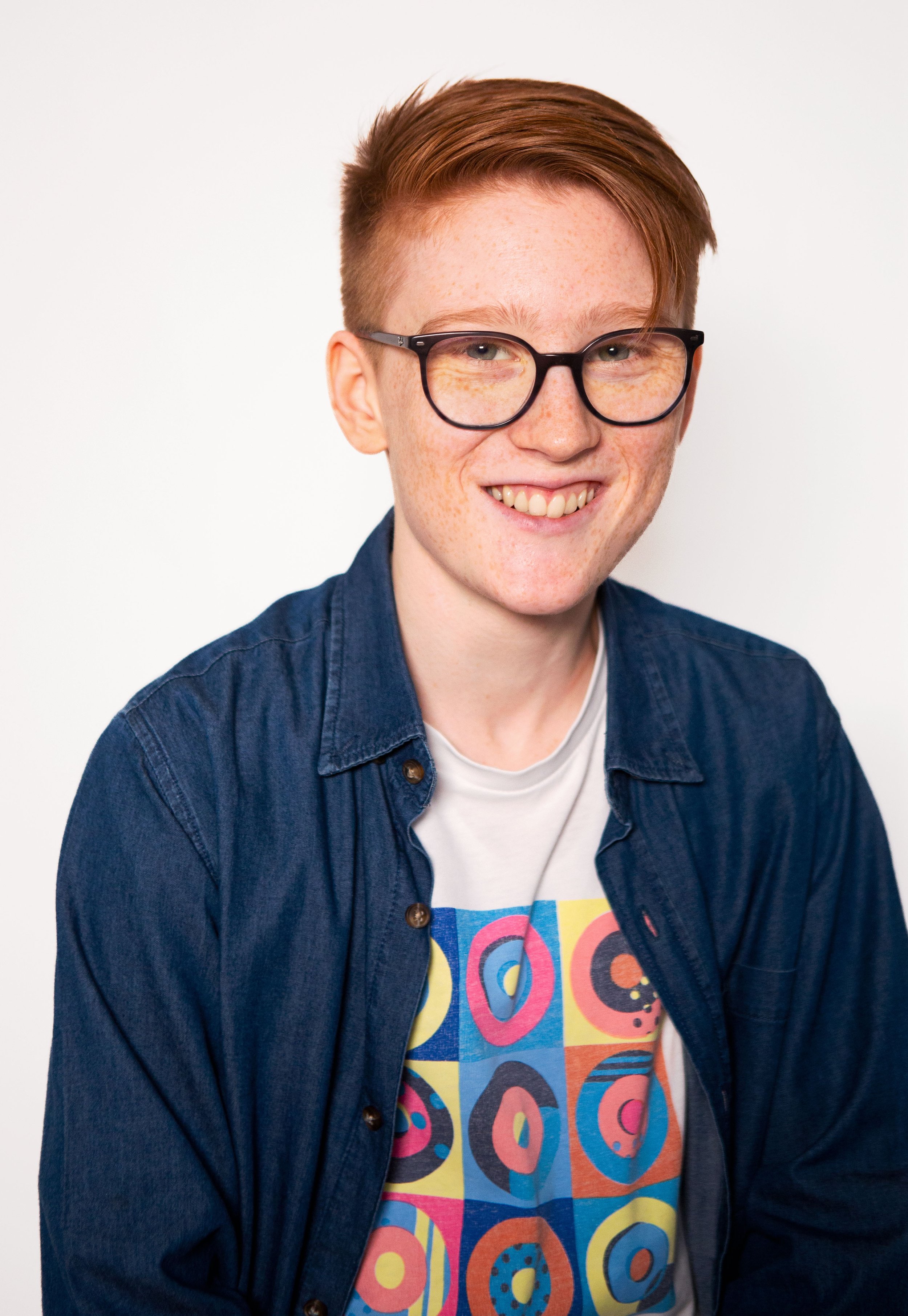 A young person with red hair, freckles, and black glasses smiling, wearing a navy blue button-up shirt over a colourful graphic T-shirt with geometric patterns, against a plain white background.