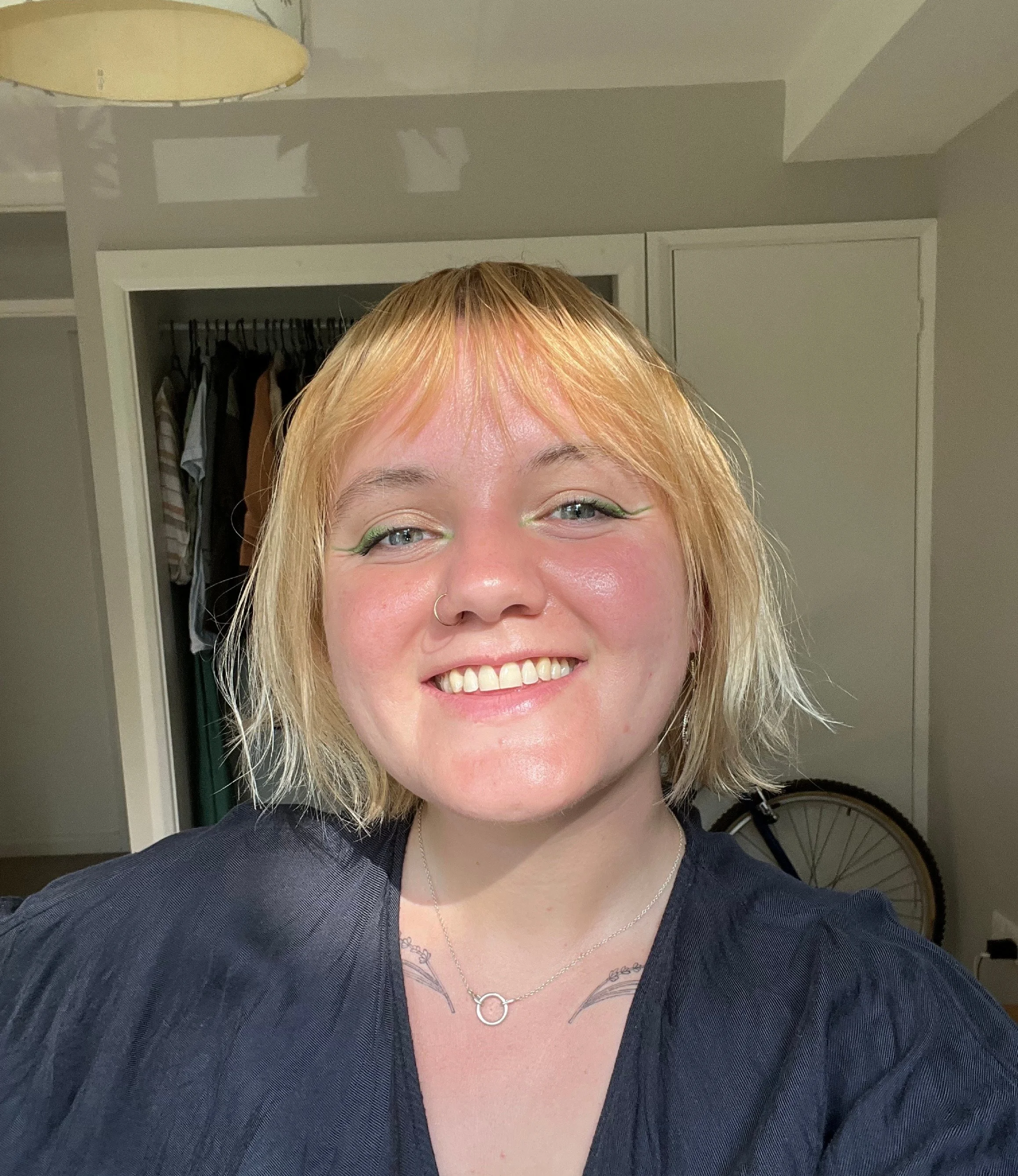 A woman with short, blonde hair and light blue eyes smiling at the camera in a room with a closet and a bicycle in the background.