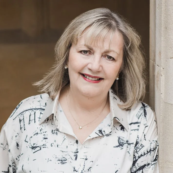 A middle-aged woman with shoulder-length blonde hair, wearing a white shirt with black abstract patterns, smiling, standing next to a stone column indoors.