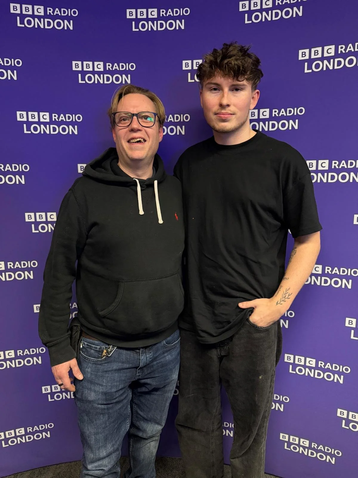 Two men standing in front of a BBC Radio London backdrop, smiling at the camera. One is wearing glasses, a black hoodie, and jeans; the other is in a black t-shirt and black pants.