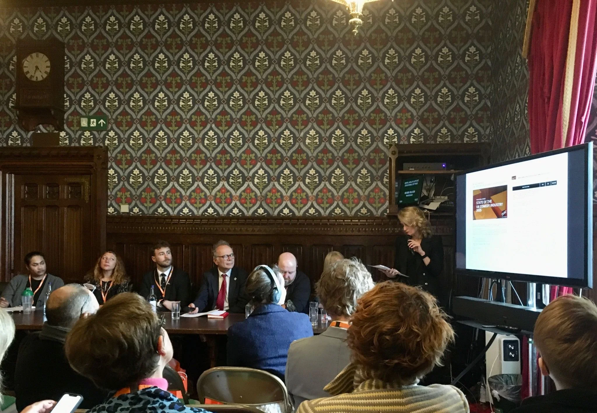 People attending a conference or meeting in a historic room with patterned wallpaper, a large screen displaying a presentation, and a woman speaking into a microphone.