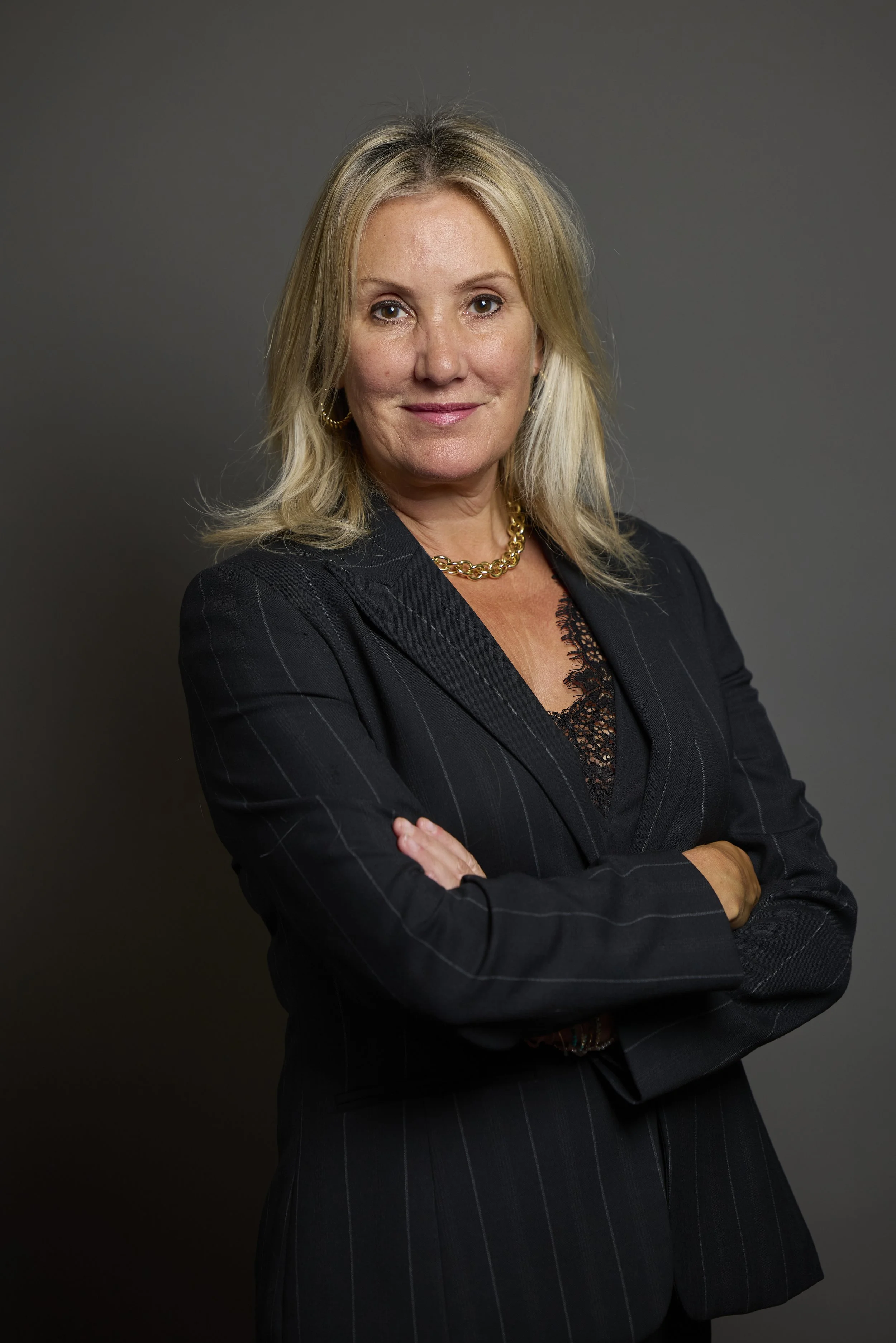 A confident woman with shoulder-length blonde hair, wearing a black pinstripe blazer over a black lace top, gold jewelry, and standing with arms crossed against a plain gray background.