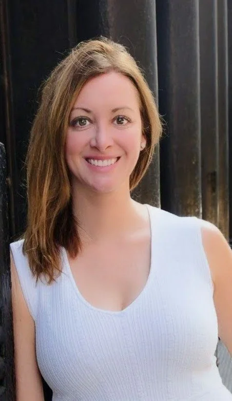 A woman with shoulder-length light brown hair, smiling, wearing a white sleeveless top, standing outdoors against a black metal fence.