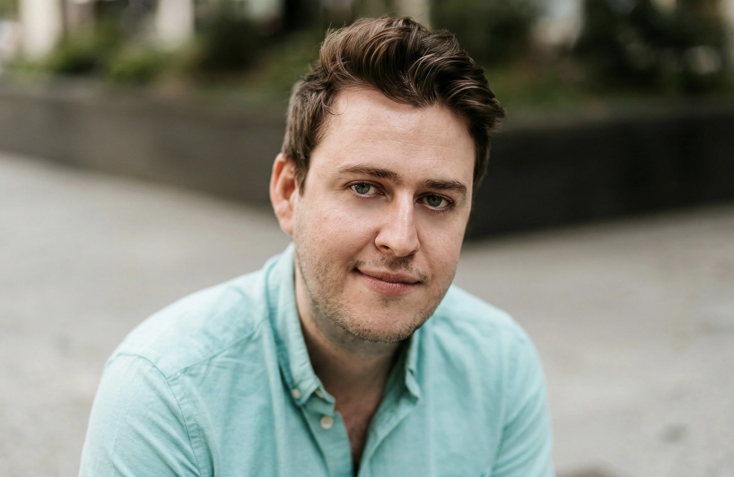 A young man with short brown hair, blue eyes, and light stubble, wearing a light blue shirt, outdoors in front of a blurred background.