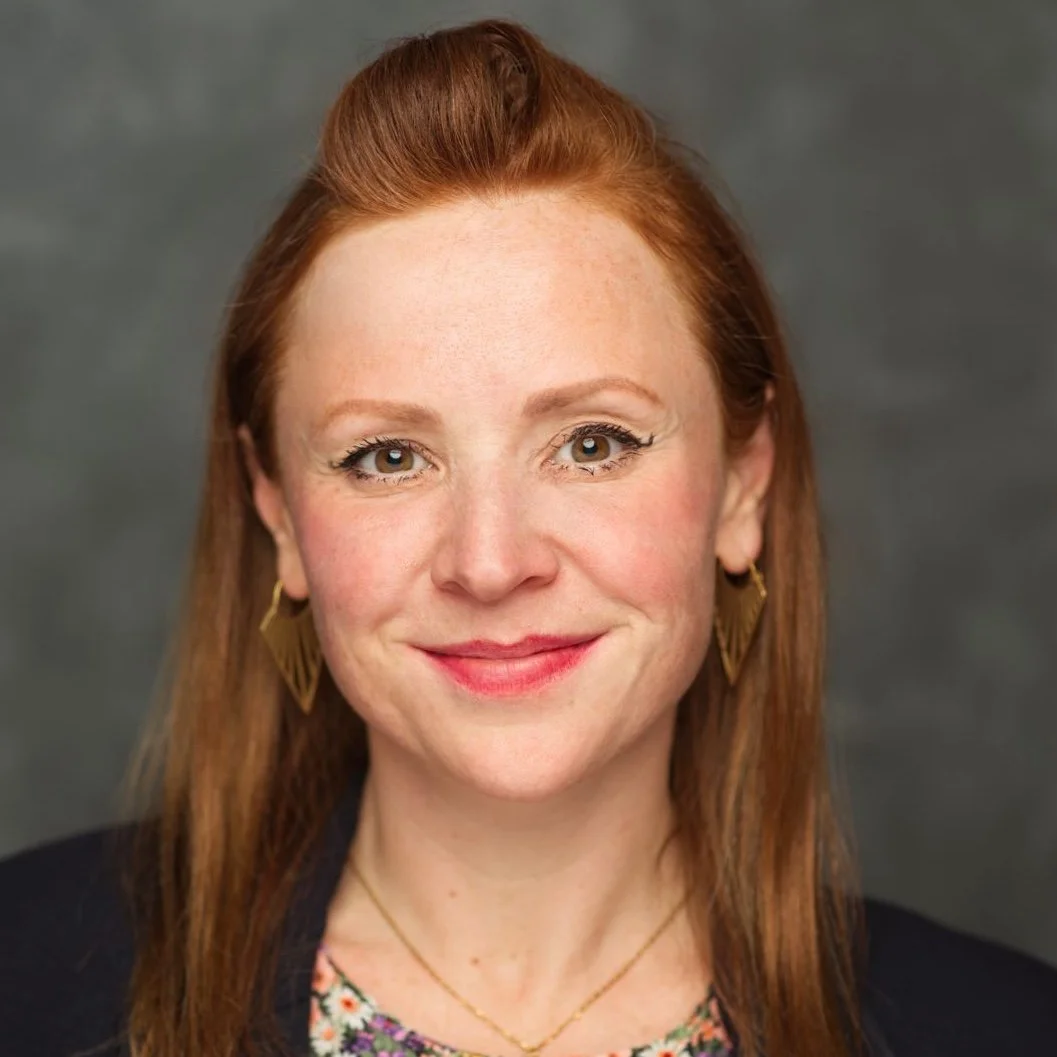 Headshot of a woman with long red hair, wearing gold earrings and a floral top, smiling against a gray background.