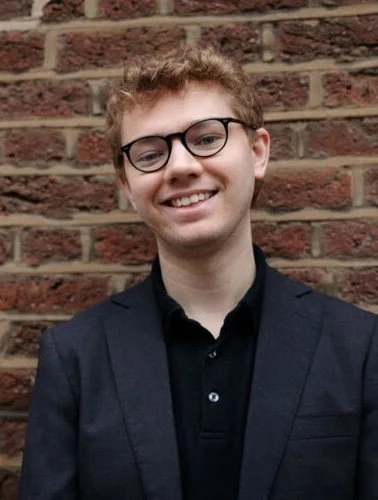 Young man with glasses, smiling, wearing a dark blazer over a black shirt, standing in front of a brick wall.
