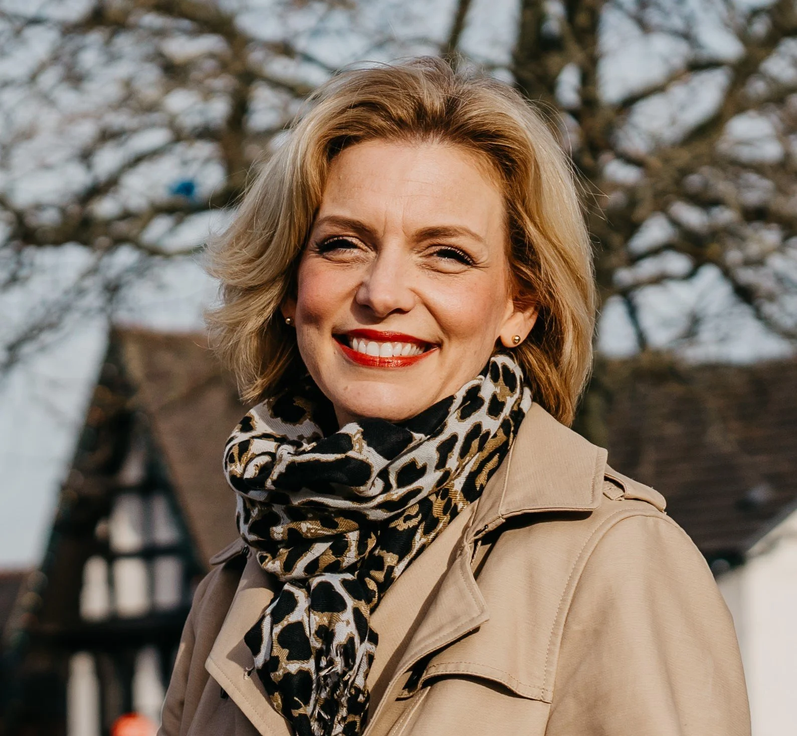 Smiling woman with short blonde hair wearing a beige coat and leopard print scarf outdoors on a sunny day, with trees and a house in the background.