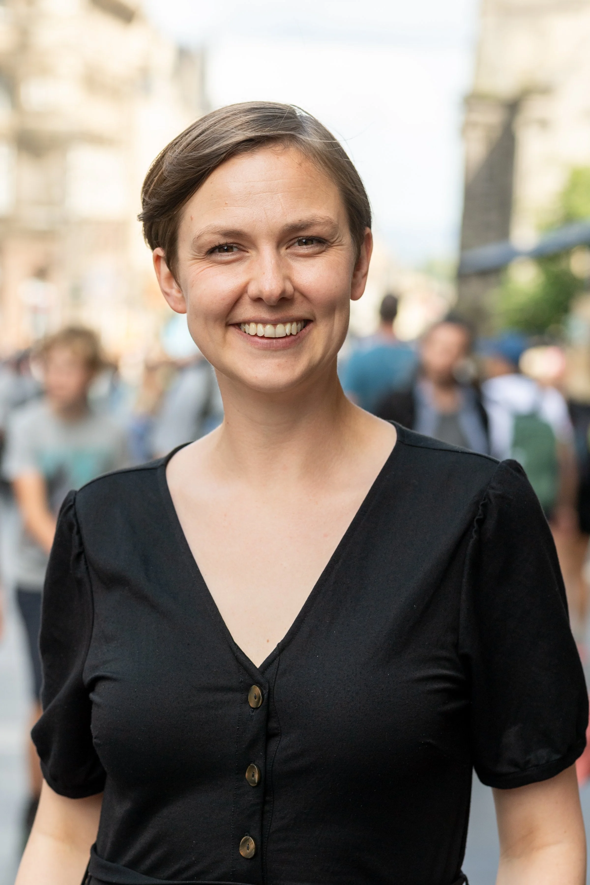 A smiling woman standing outdoors among a blurred crowd on a city street.