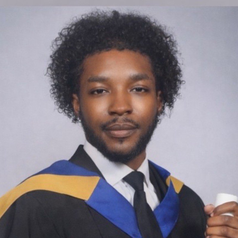 Graduation portrait of a young man with curly hair and beard wearing a gown with blue, black, and gold accents, holding a diploma.