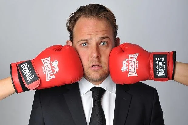 Man in a suit wearing red boxing gloves, holding them up to his head in a boxing pose, with a serious facial expression.
