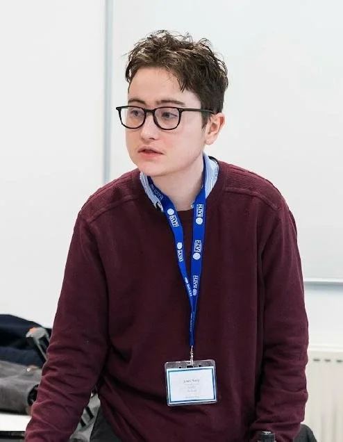 A young man with curly brown hair wearing glasses and a maroon sweater, standing in a classroom or conference room with a whiteboard in the background, looking off to the side with a serious expression.