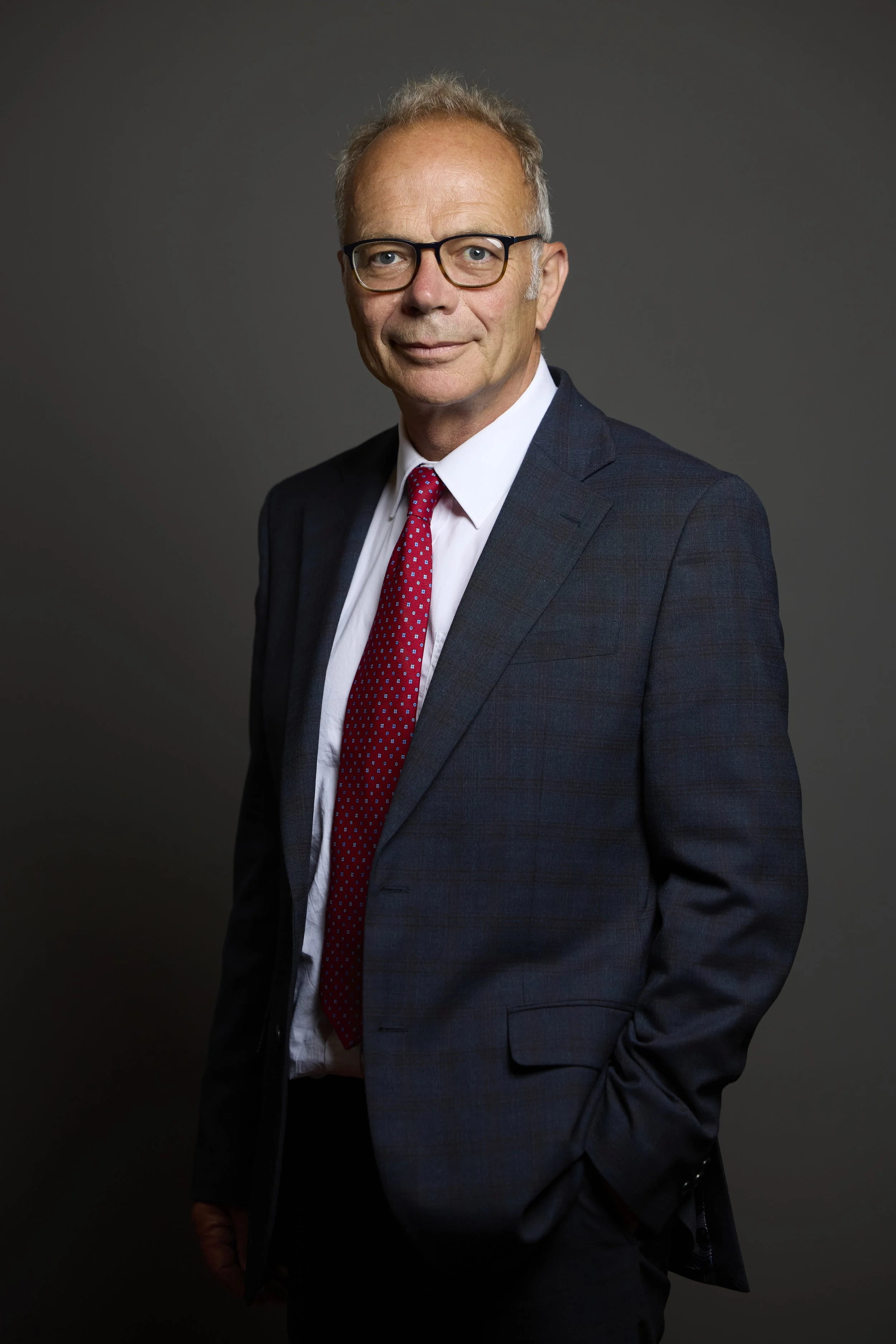 A middle-aged man in a dark suit, white shirt, red tie, and glasses posing against a plain background.