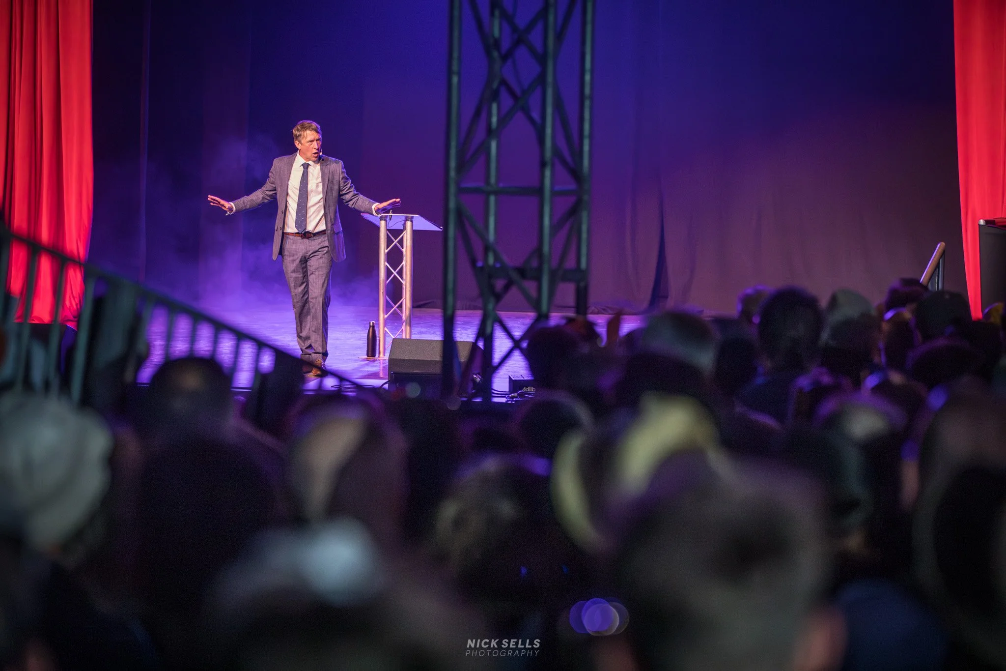 Male speaker on stage at a conference or event, gesturing with his hands, with an audience watching, purple and red lighting, and stage equipment visible.