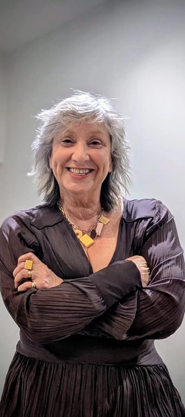 Smiling older woman with gray hair, wearing a dark shiny dress and gold jewelry, crossing her arms against a plain background.