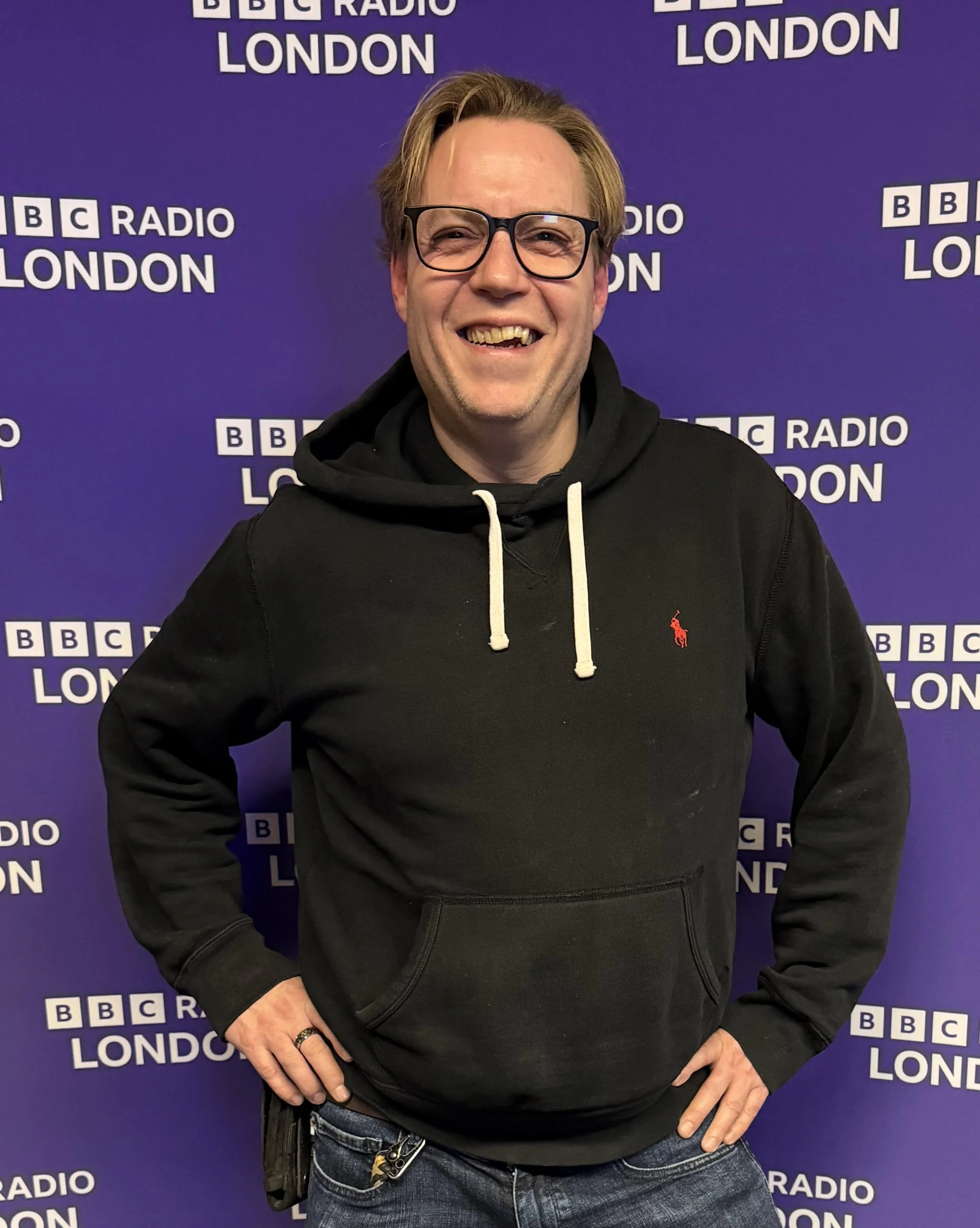 A man wearing glasses, a black hoodie with white drawstrings, and blue jeans, standing in front of a purple background with the BBC Radio London logo repeatedly printed on it. He is smiling with one hand on his hip.