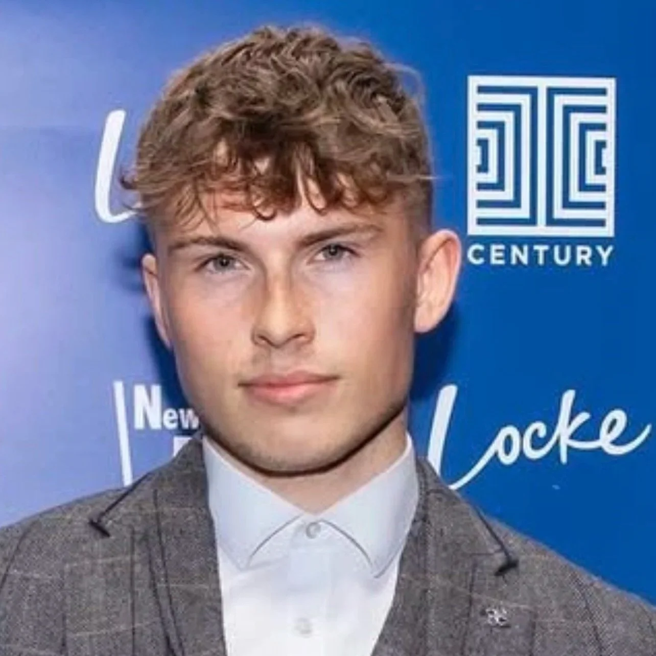 A young man with curly light brown hair, wearing a gray checkered suit and white shirt, posing in front of a blue background with logos and text.