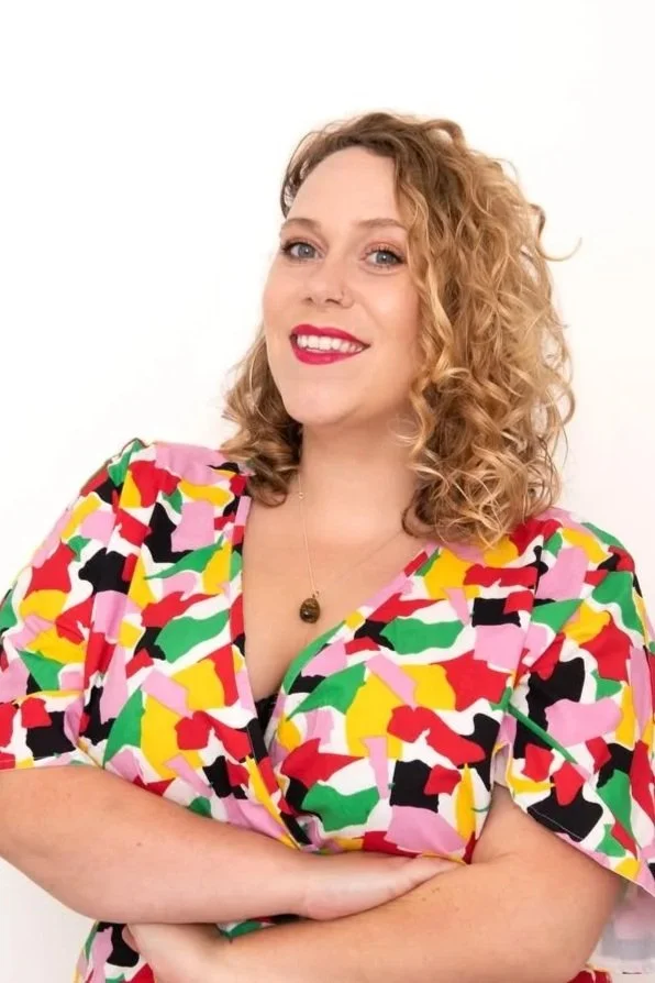 A woman with curly, shoulder-length blonde hair, wearing bright lipstick and a colorful, patterned dress with short sleeves, smiling against a plain white background.