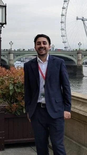 A man in a dark suit and white shirt standing outdoors by the river, with London's London Eye and a bridge in the background.