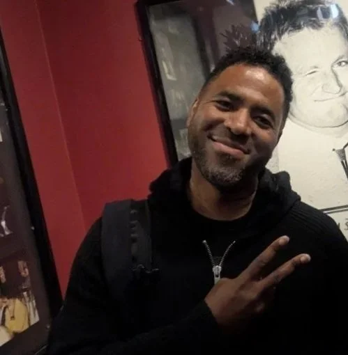 A man with short curly hair and a beard smiling and making a peace sign with his hand, standing indoors near a wall with a large black-and-white drawing of a person's face.
