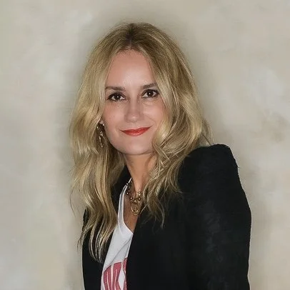 A woman with blonde hair wearing a black blazer and white t-shirt, smiling at the camera