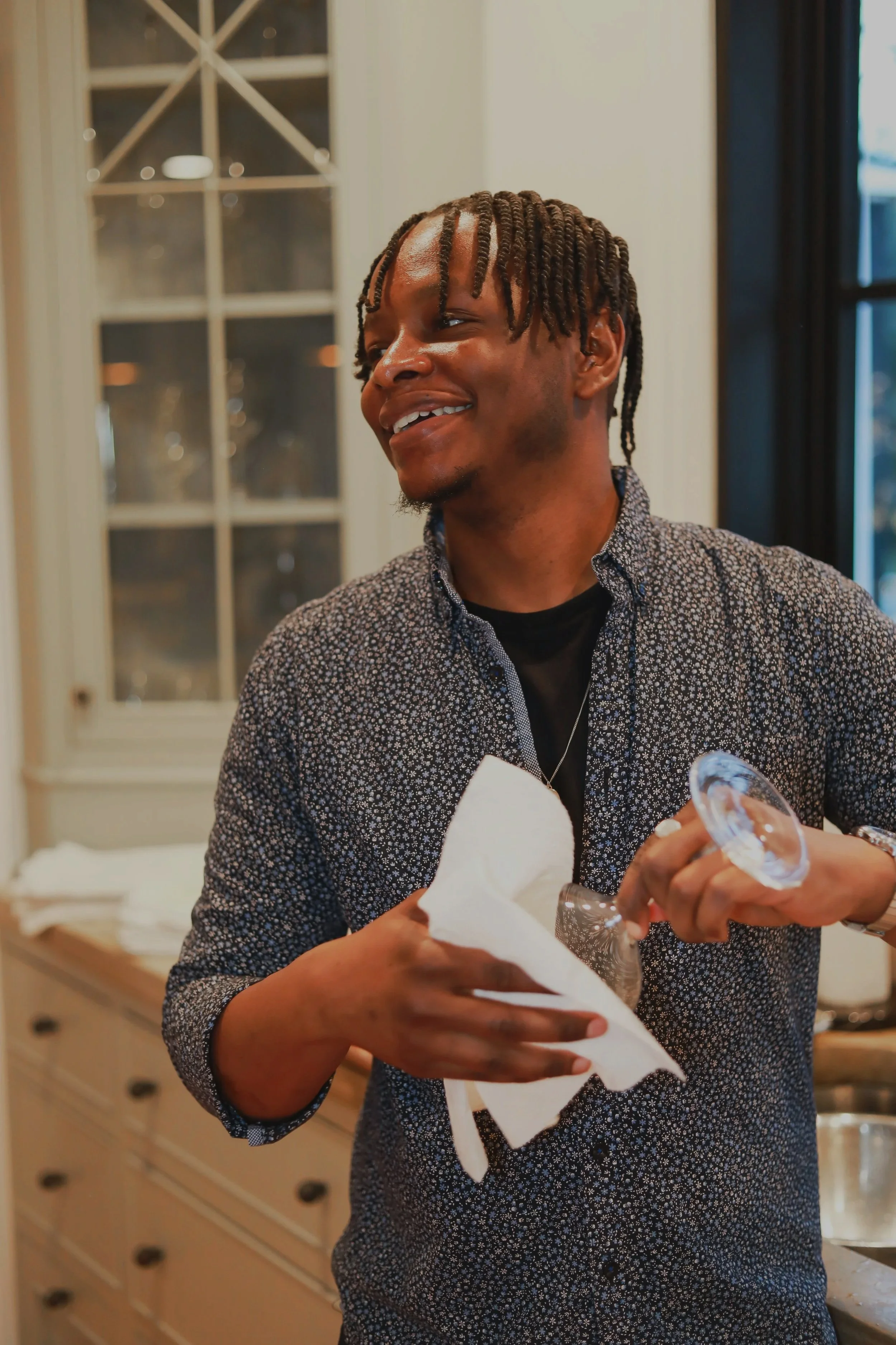 A smiling man with twisted hair in a patterned shirt holding a wine glass and a napkin in a kitchen with light-colored cabinets and a window.