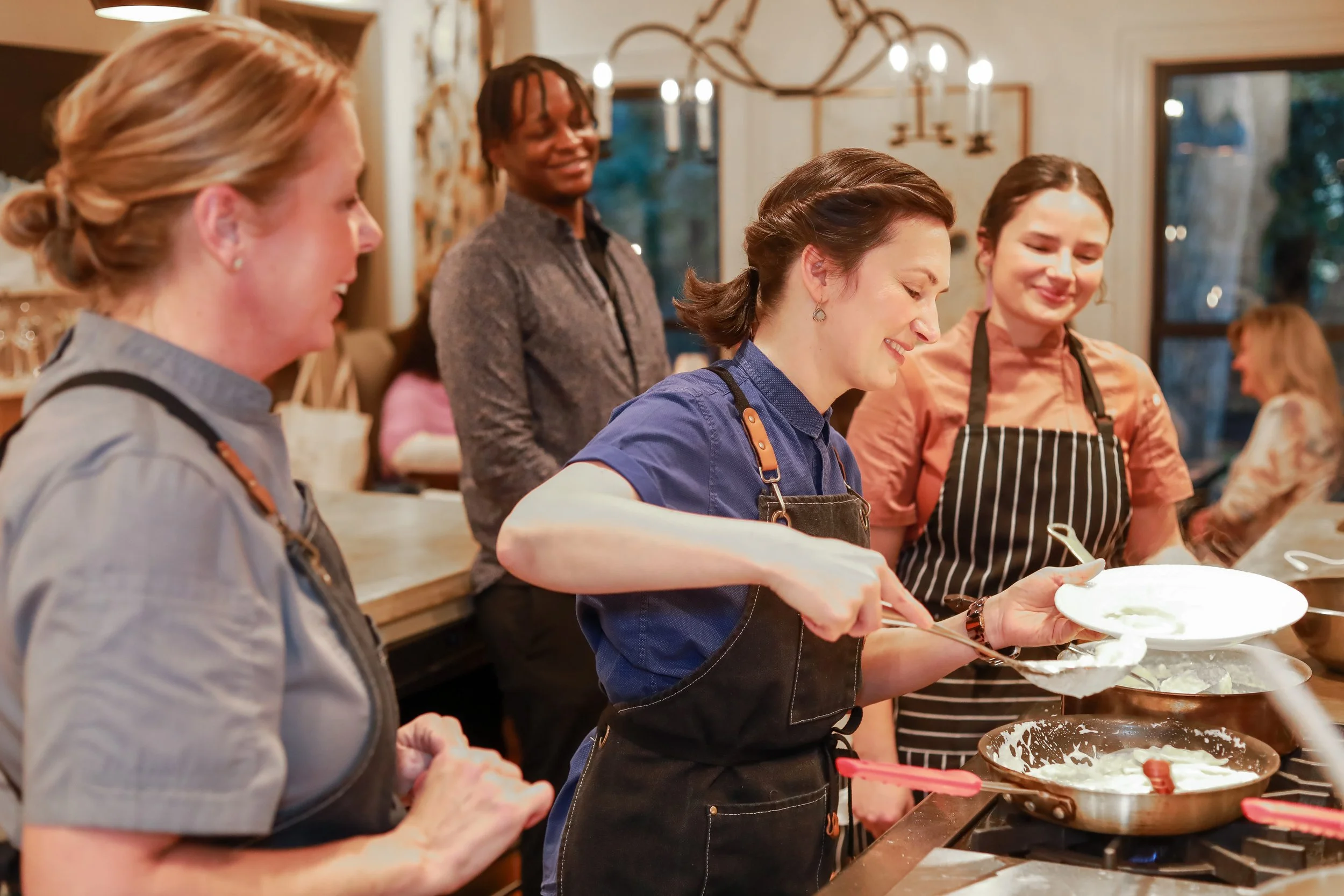 A group of people in a kitchen cooking together, with one woman scooping batter into a bowl, while others watch and smile.