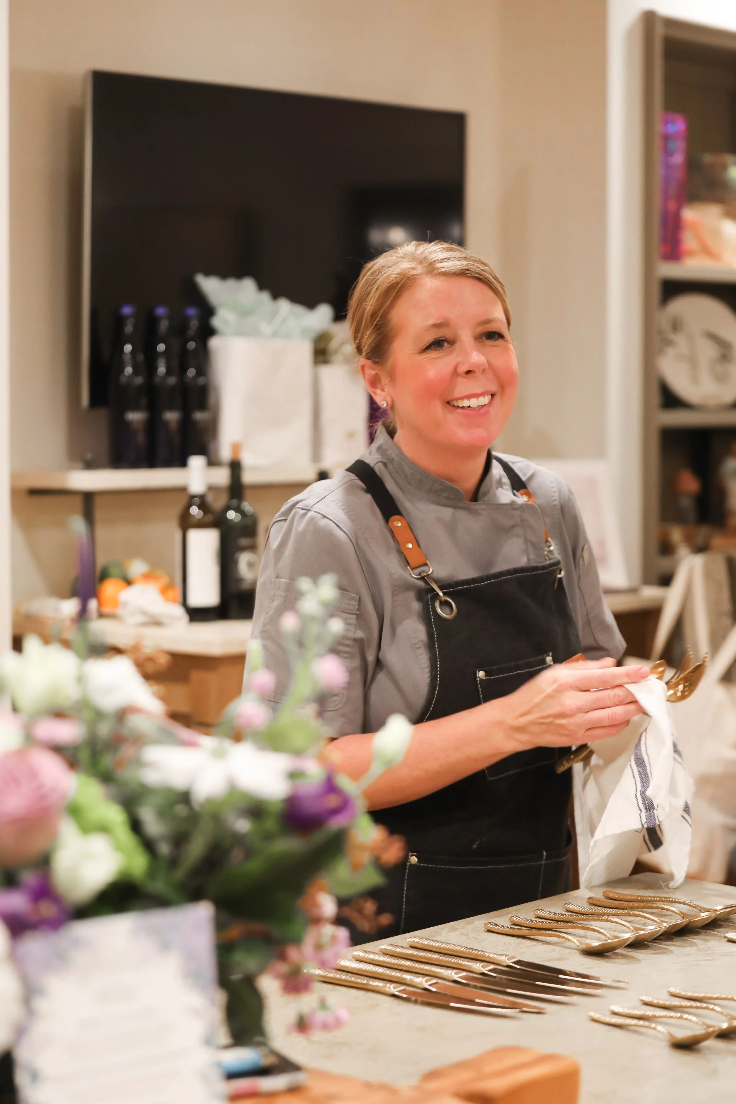 A smiling woman in a gray shirt and black apron standing behind a table with neatly arranged gold spoons, holding a white cloth, with a floral arrangement in the foreground, shelves and a television in the background.