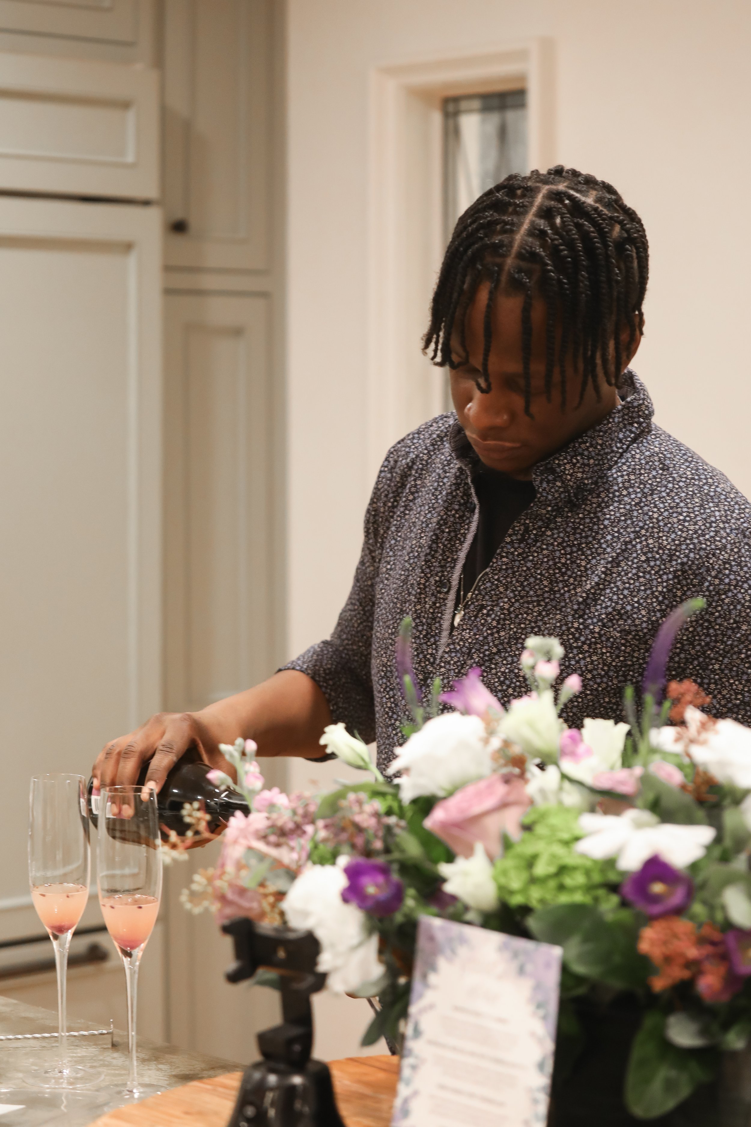 A young man with twisted hair is pouring champagne into glasses, with a floral centerpiece in the foreground.