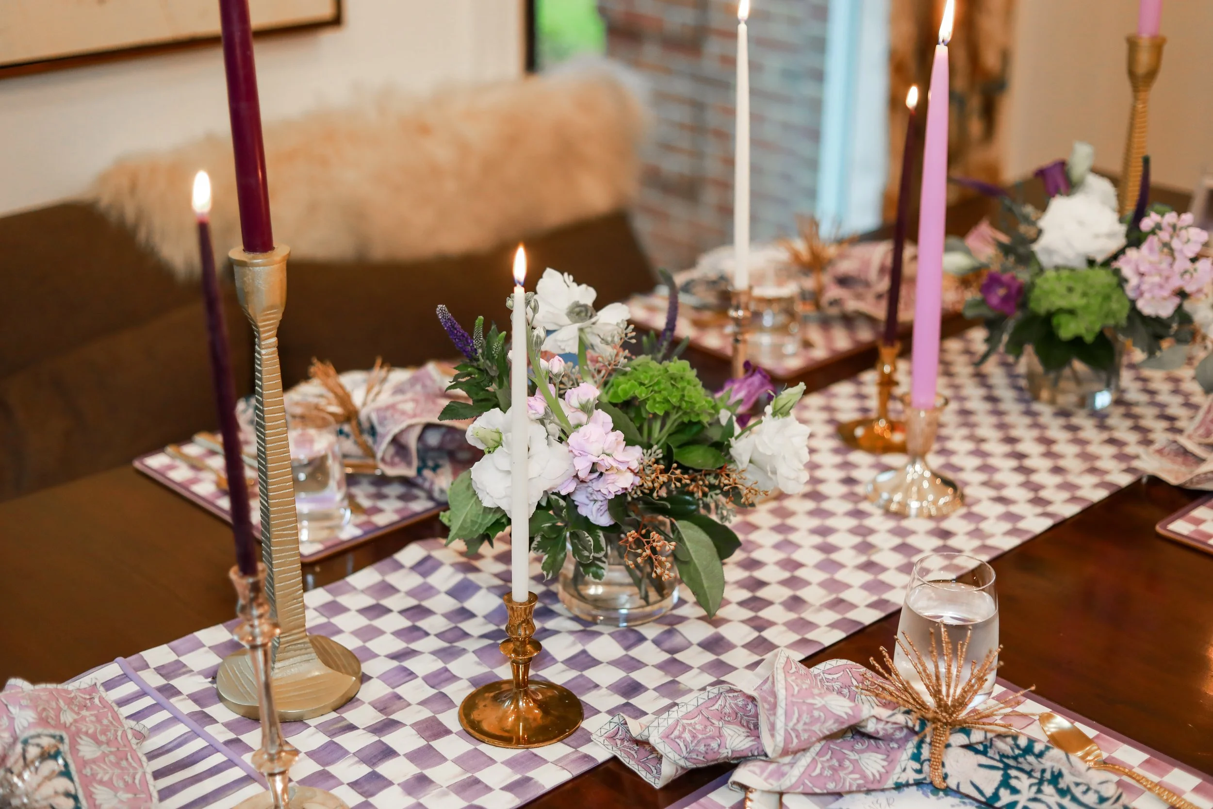 A dining table decorated with multiple pink, purple, white candles in ornate candleholders, a floral centerpiece with pink and white flowers and green foliage, and pink and purple table settings with cloth napkins and patterned placemats.