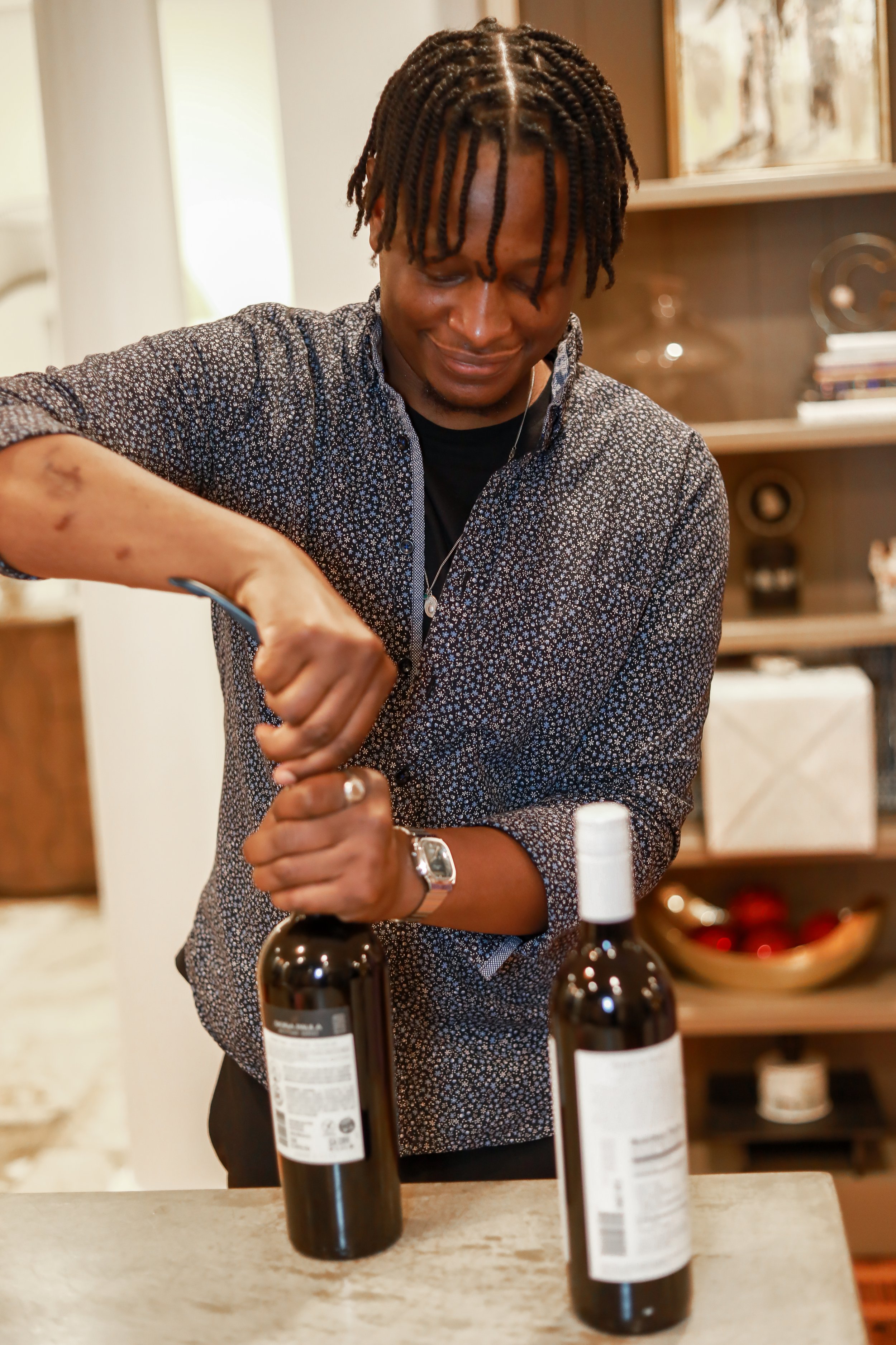 A young man with twisted hair and a patterned button-up shirt looks down and smiles as he opens a wine bottle, surrounded by three bottles on a light-colored countertop, in a room with wooden shelves and framed pictures.