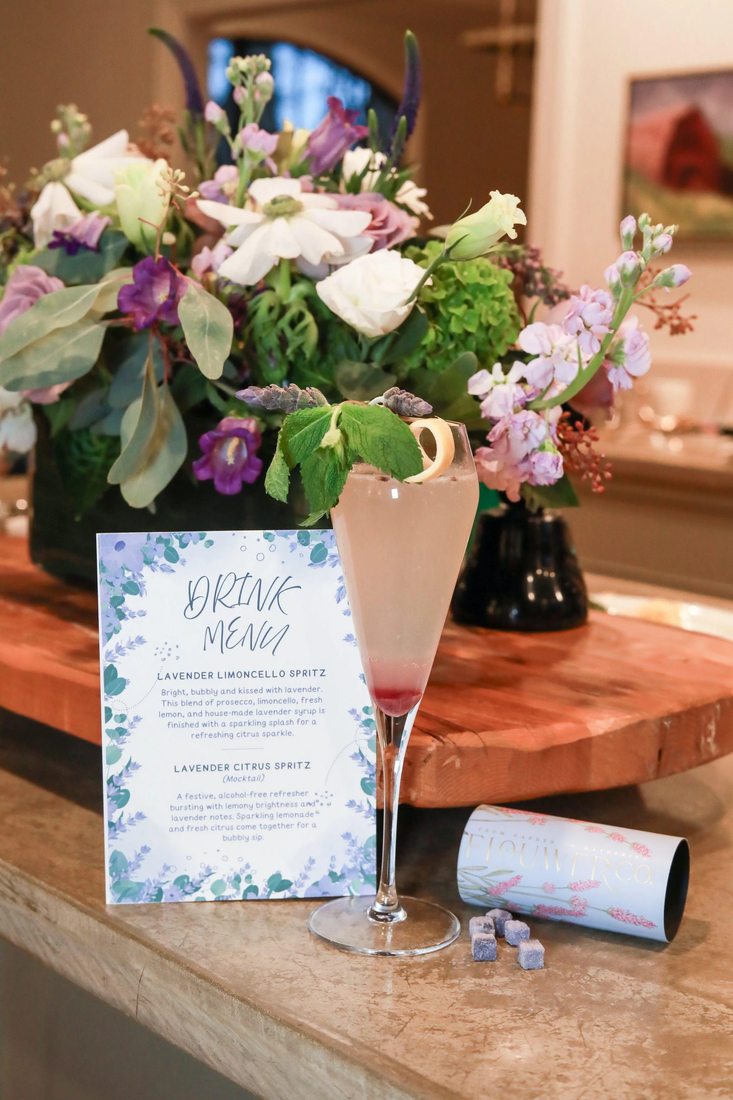 A tall glass of lavender lemonade spritz garnished with a lemon twist and lavender sprig, placed on a wooden table next to a floral-themed drink menu and a lavender flower packaging.