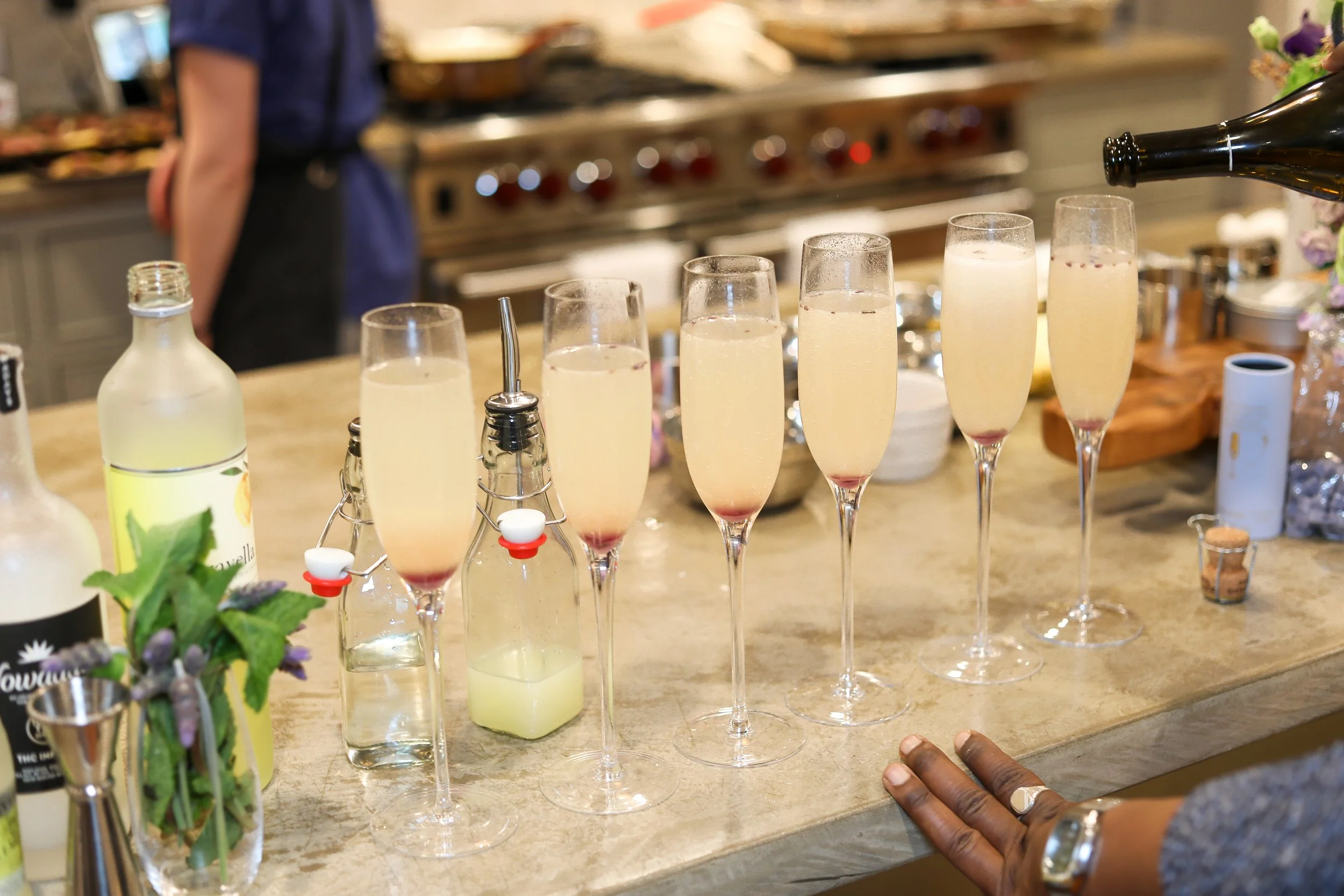 A row of champagne glasses filled with a champagne spritz drink and fruit, on a kitchen counter with bottles and utensils in the background.