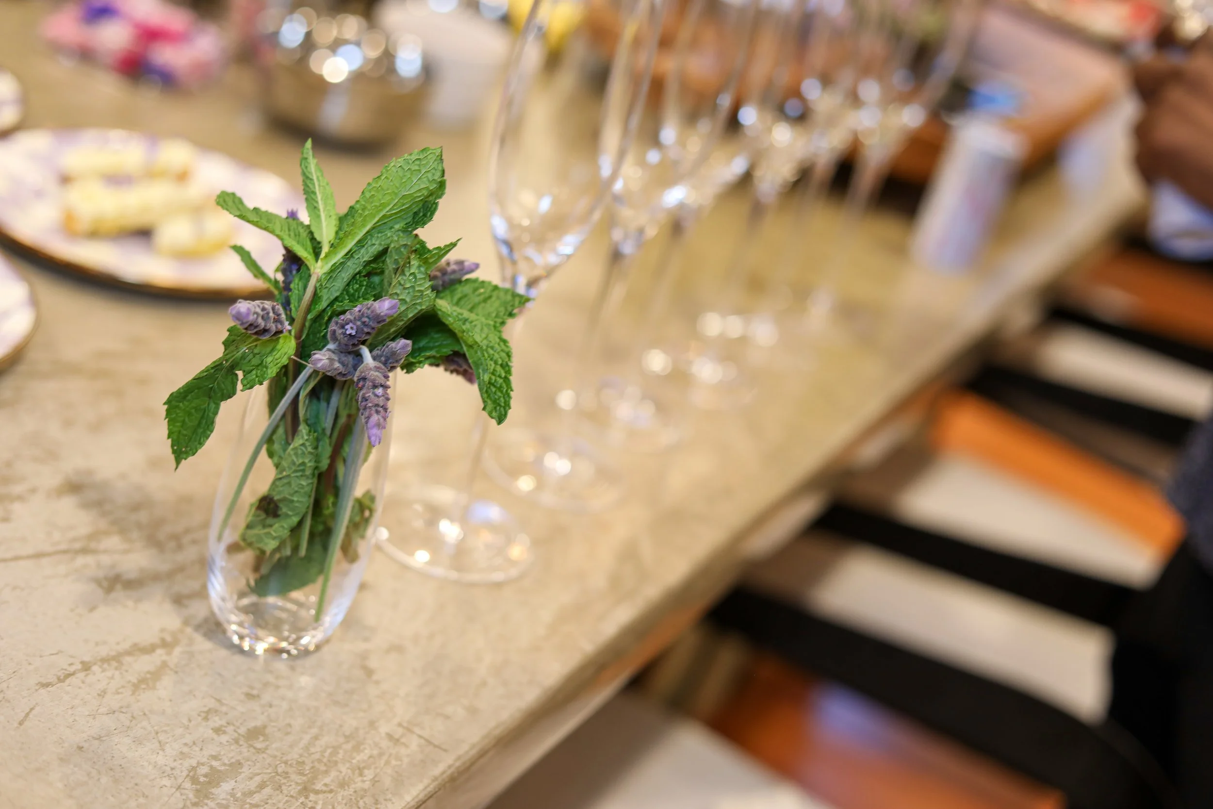 A small glass vase with a bouquet of fresh green mint and lavender sprigs on a beige countertop, with blurred champagne glasses and party supplies in the background.