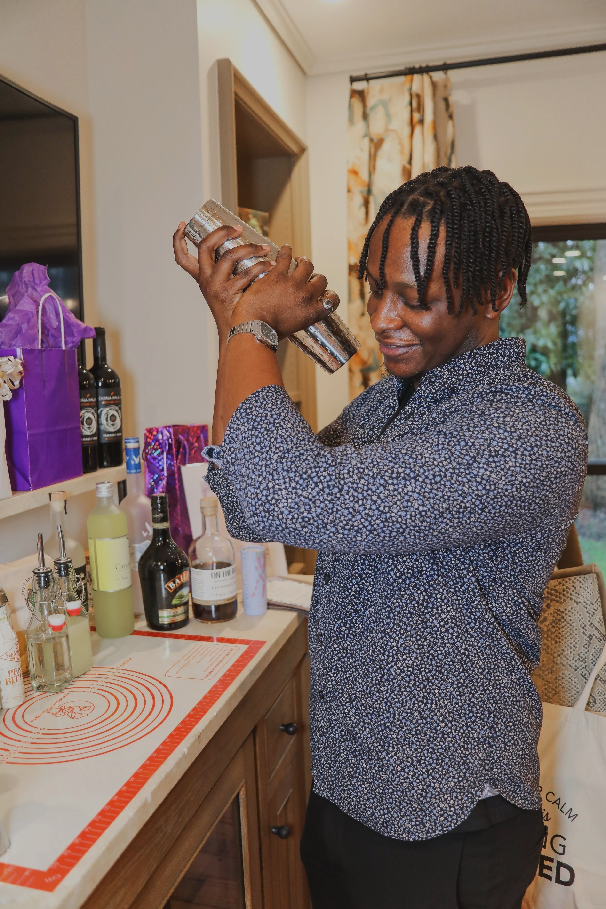 A young man with twisted hair is wearing a patterned shirt and holding a cocktail shaker near his face, appearing to shake or prepare a drink inside a kitchen with a window, curtain, and various bottles and bar accessories on the counter.
