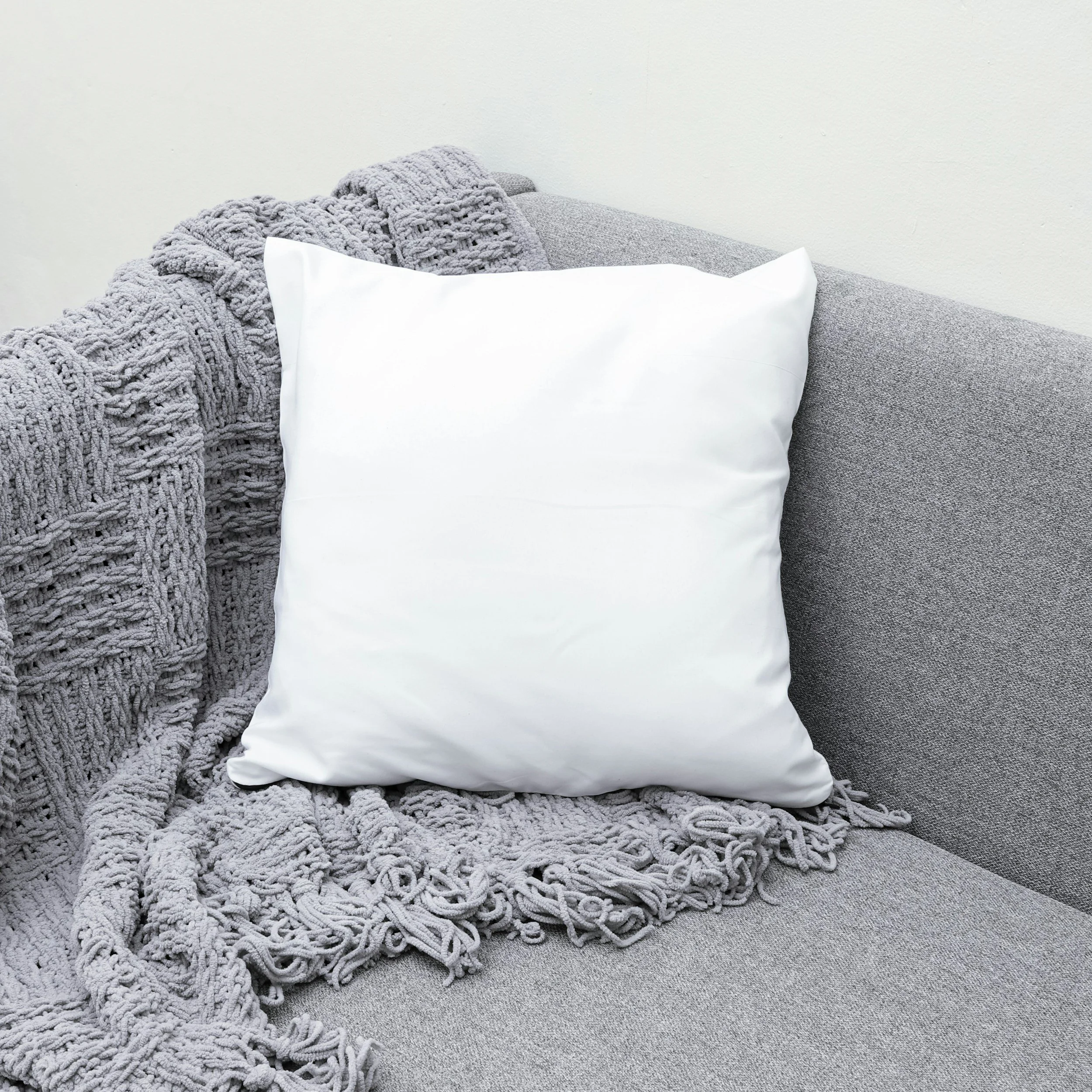 A grey couch with a chunky knit blanket draped over the armrest and back, a white pillow, and a textured grey pillow, set against a plain white wall.