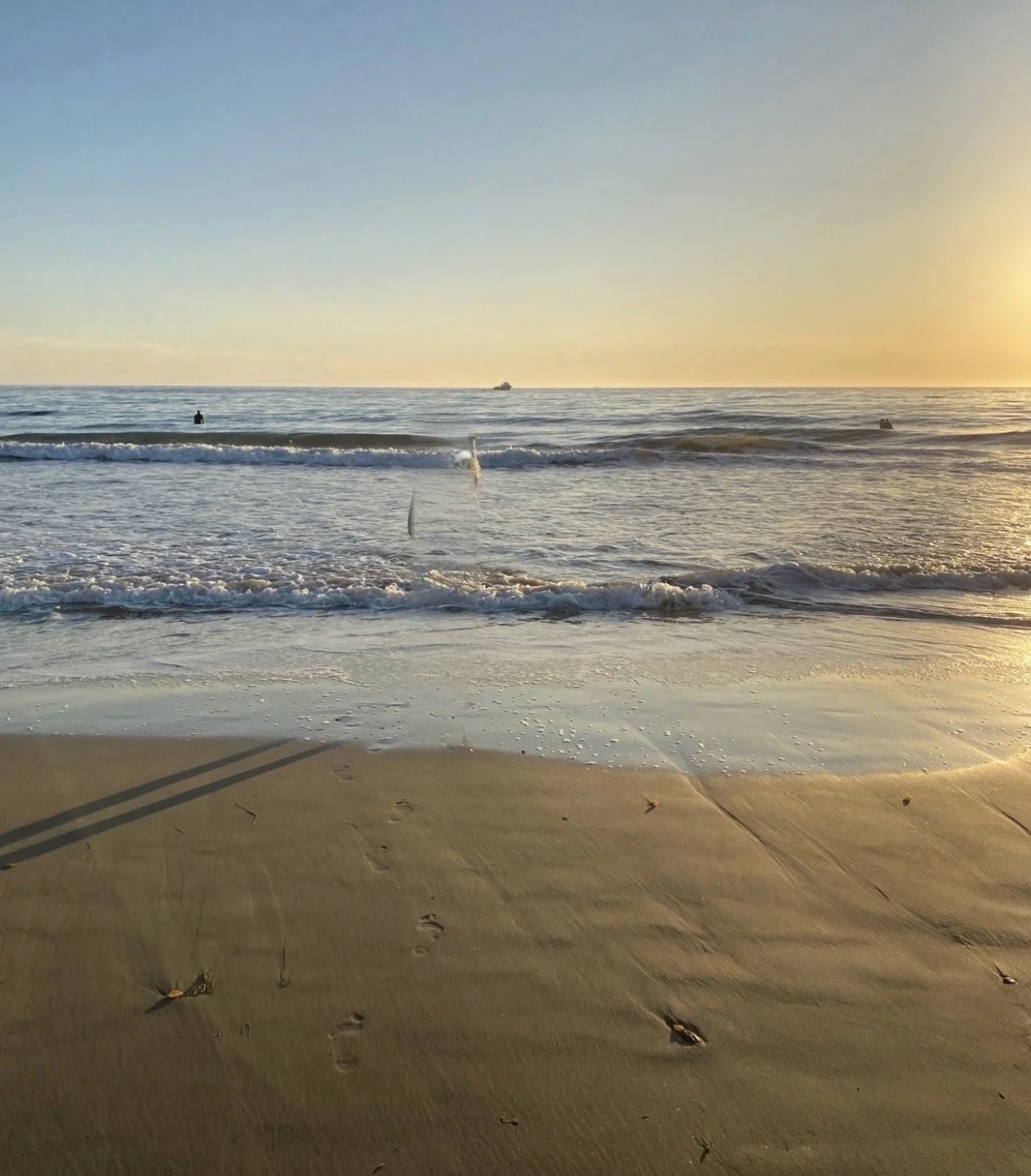 Sunset at the beach with footprints in the sand, small waves, and a ship on the horizon.
