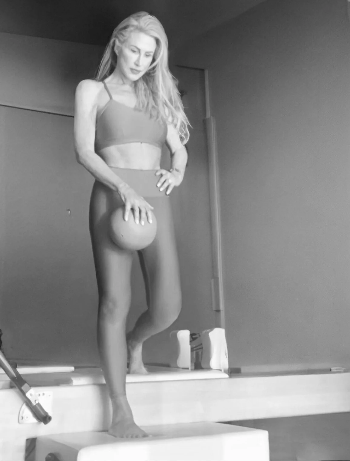 A woman with long blonde hair wearing a sports bra and leggings, standing on a platform, holding a ball in her hand, looking down thoughtfully.