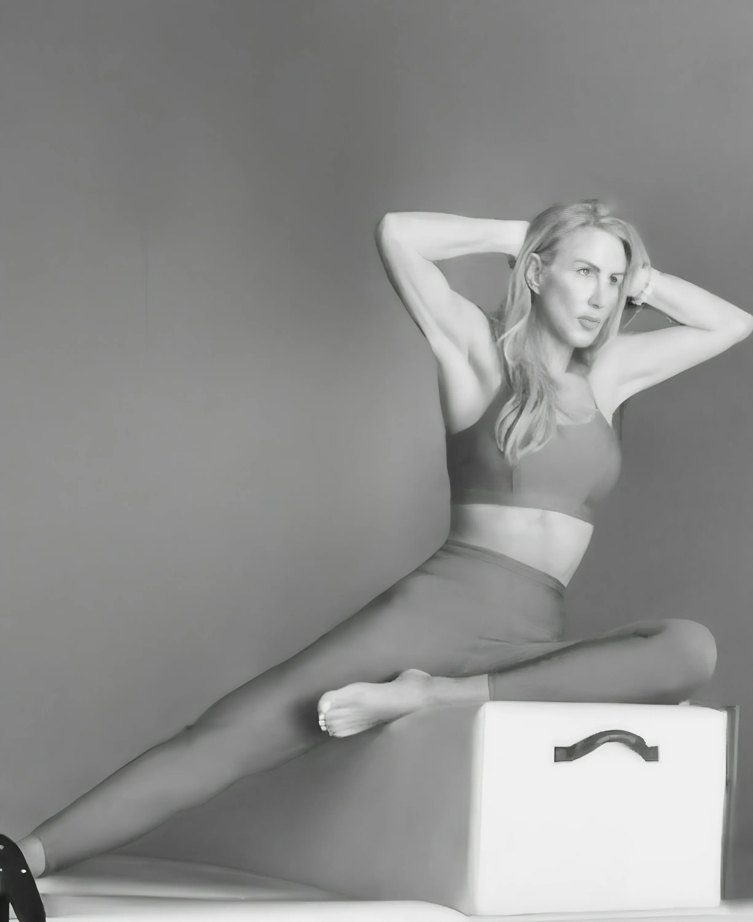 A woman with long hair in a sports bra and leggings sitting on a white box, stretching with hands behind her head, against a plain background.