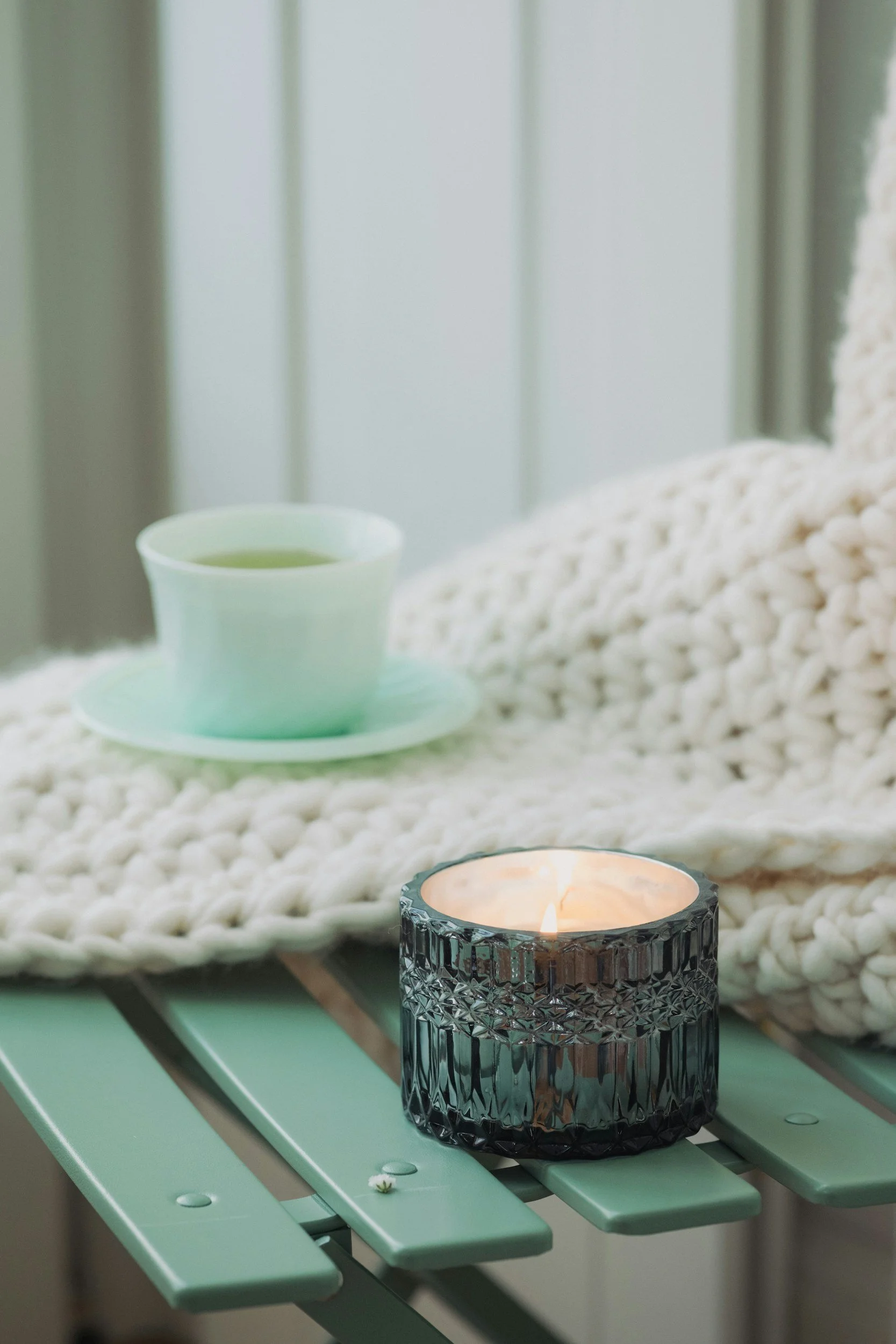 A mint green outdoor table with a lit candle in a textured black glass holder, a white knit blanket, and a white cup with a saucer in front of a paneled wall.