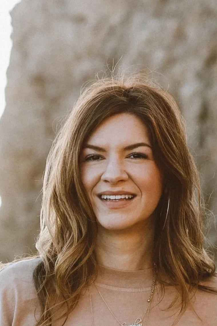 Close-up of a woman with wavy brown hair smiling outdoors with a blurred neutral background.