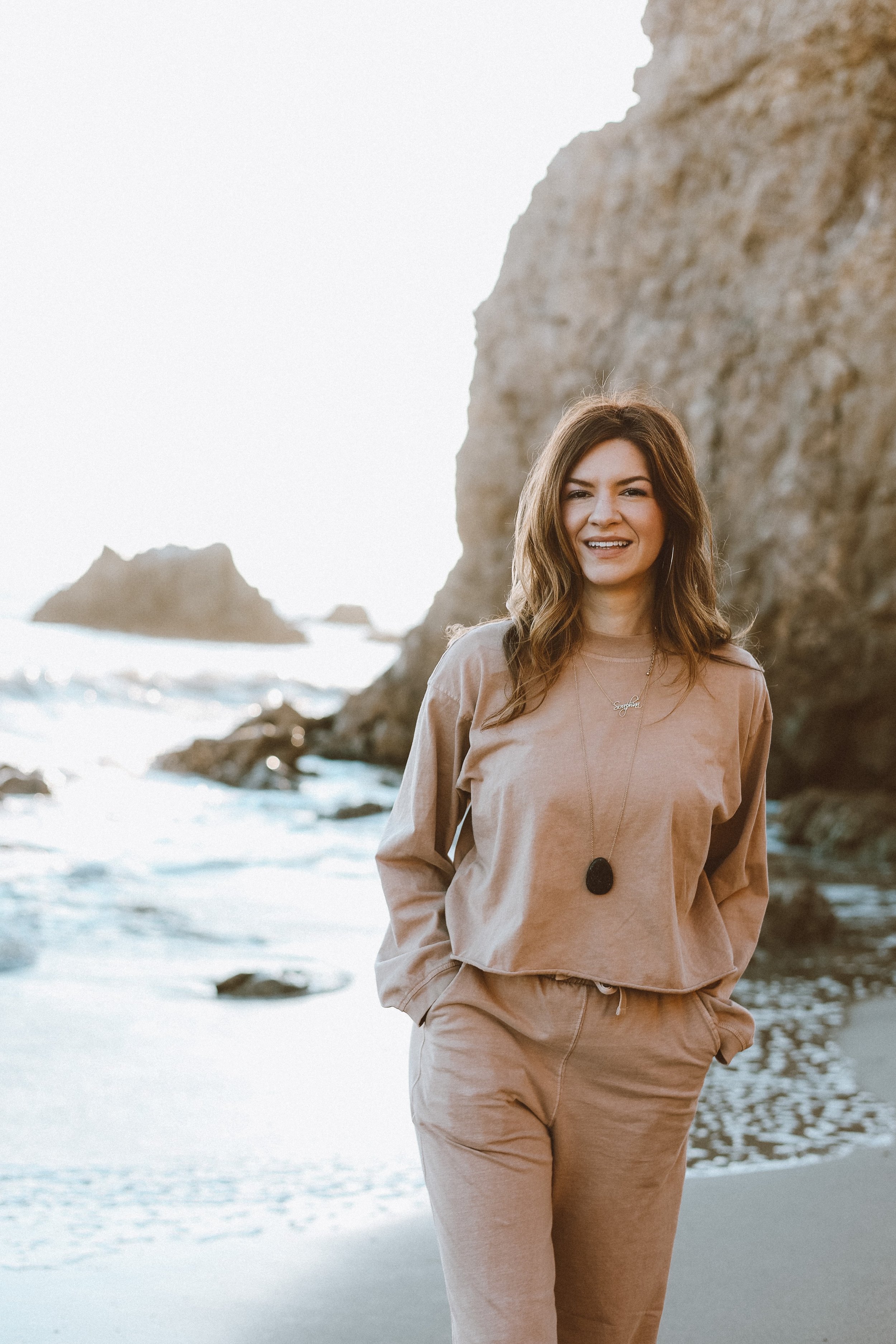 Woman with brown hair smiling at the beach near large rocks and cliffs, wearing beige loungewear and jewelry.