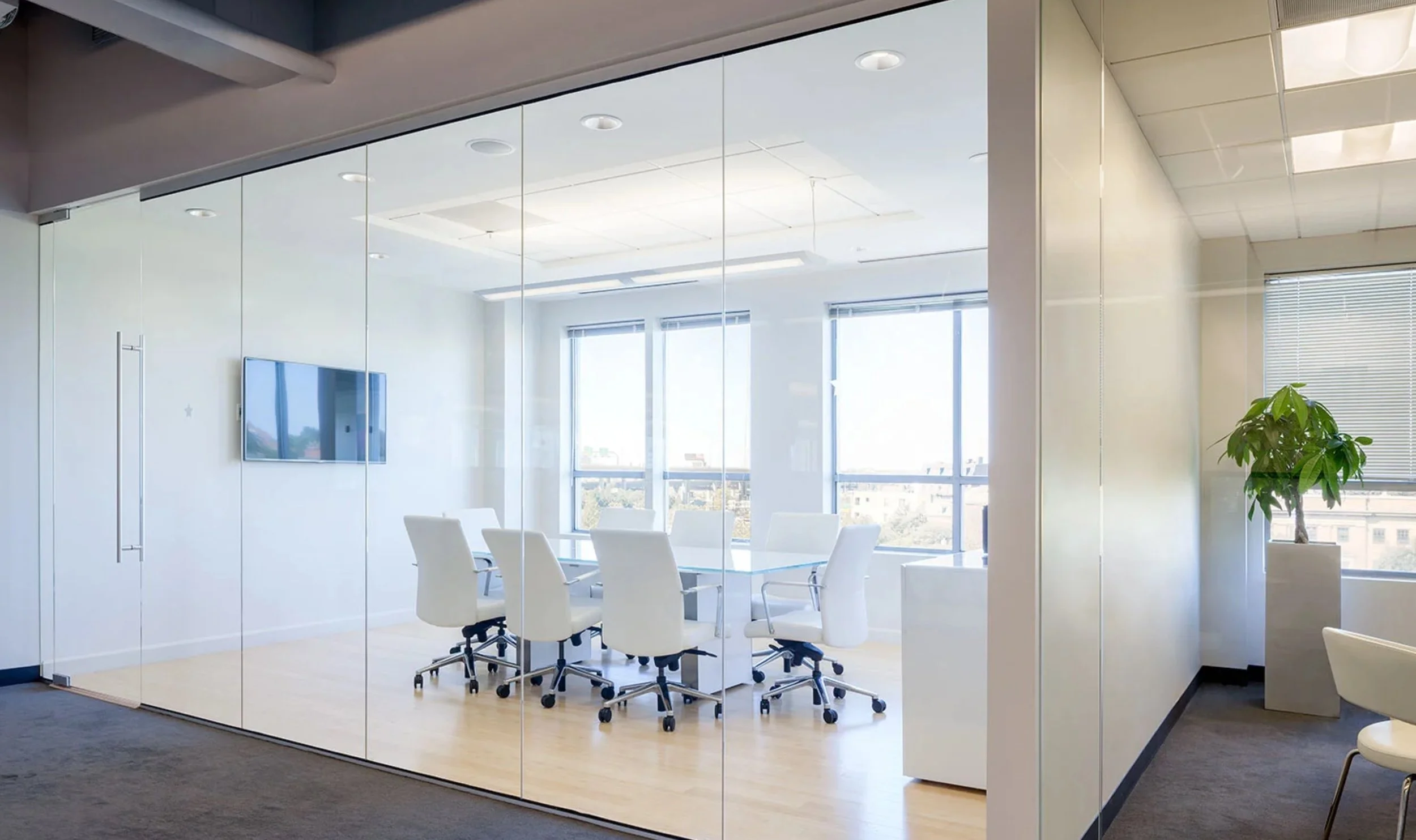 Modern conference room with a white table, six white chairs, large windows, a wall-mounted TV, and a potted plant.