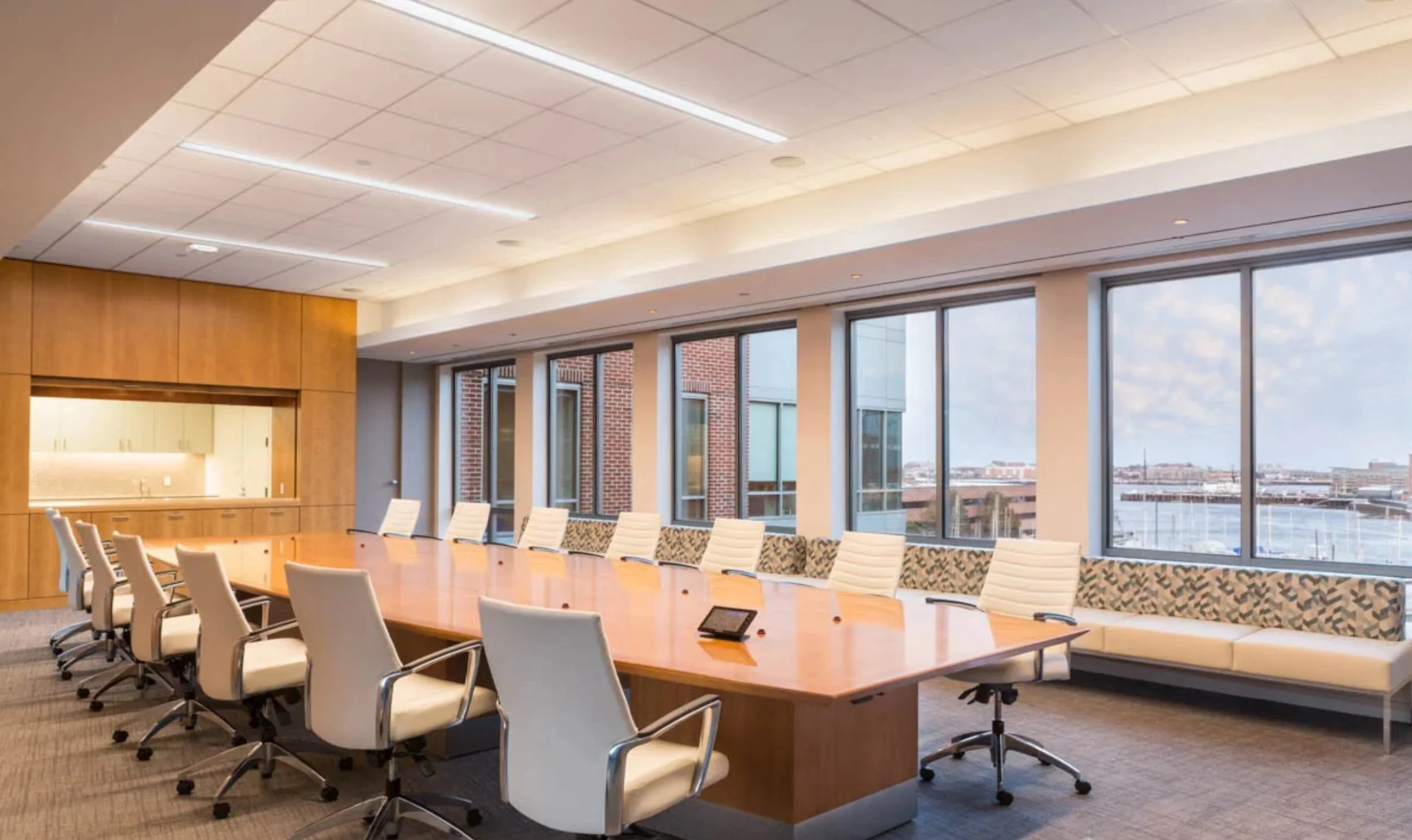 Modern conference room with a long wooden table, white chairs, large windows with city views, and a ceiling with integrated lighting.