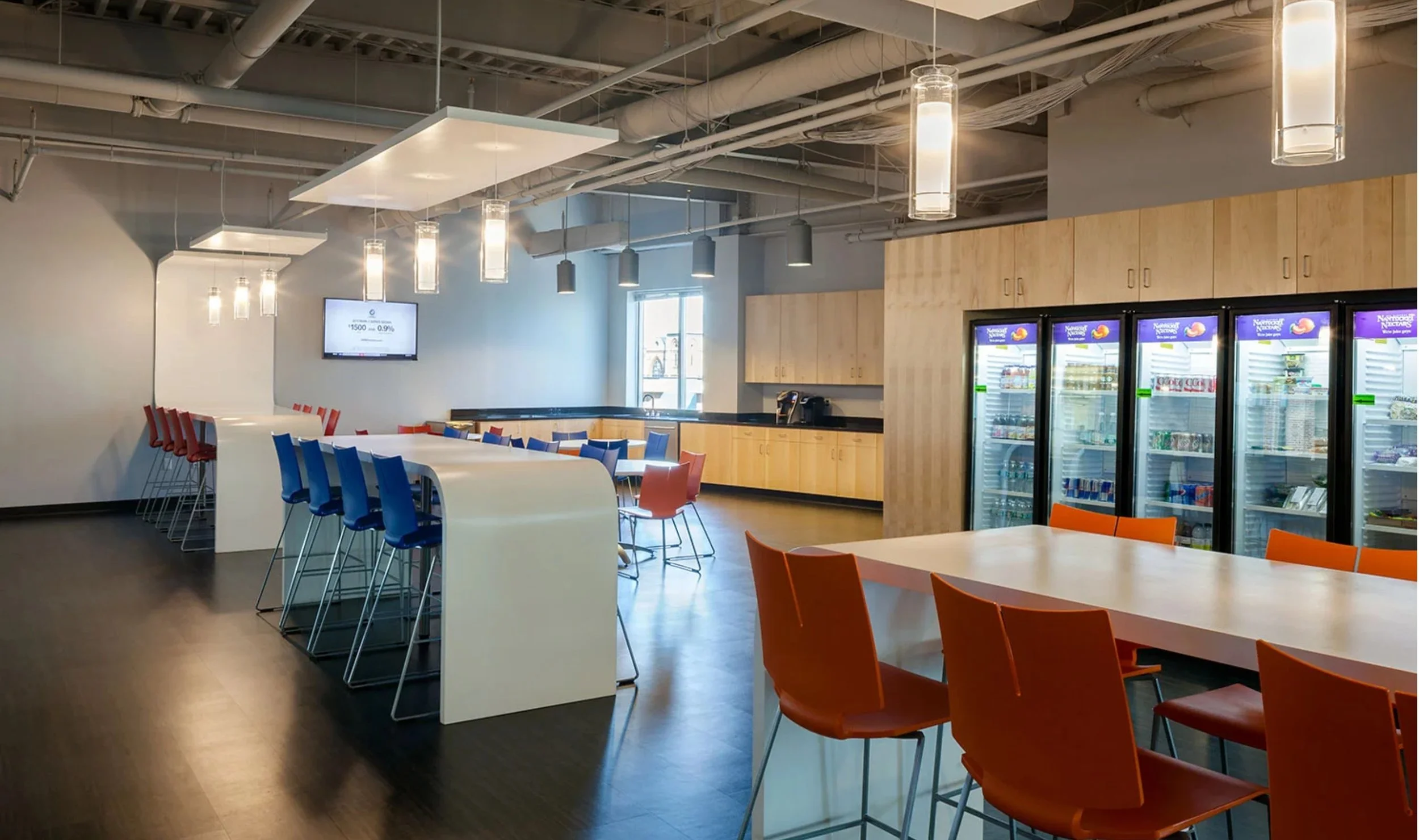 Modern break room or cafeteria with high-top tables and colorful chairs, a row of vending machines, wooden cabinets, a coffee station, a wall-mounted TV, and industrial ceiling lighting.